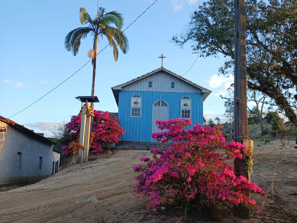 A Capela São Geraldo no alto do morro, de madeira azul, com azaleias floridas e uma palmeira ao lado, em manhã de céu limpo.